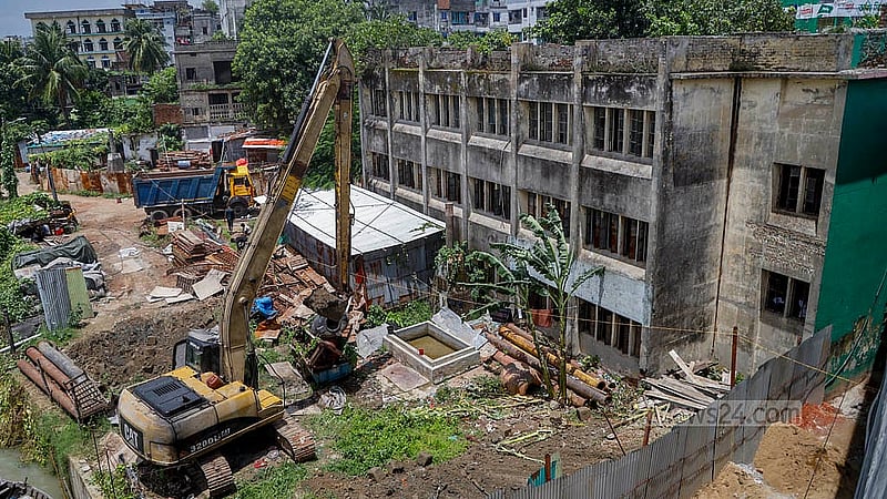 ঢাকার বছিলা সরকারি প্রাথমিক বিদ্যালয়ের গা ঘেঁষে স্কেভেটর দিয়ে মাটি খোড়ার কাজ চলছে। এই পথেই বুড়িগঙ্গা নদীর পাশ দিয়ে বানানো হবে পথচারী চলাচলের জন্য ওয়াকওয়ে