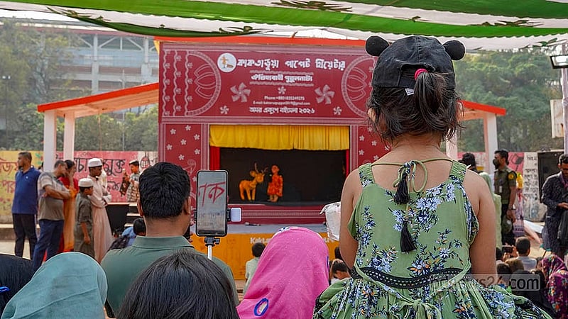 বইমেলায় এসে বাবার কাঁধে চড়ে পুতুল নাচ দেখছে এক শিশু