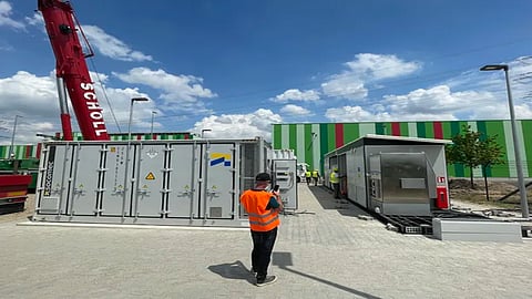 A commercial logistics and refrigerated warehouse facility in Germany with rooftop solar panels connected to two large battery energy storage containers, illustrating a 5 MW/10 MWh solar-plus-storage system that stores excess renewable electricity for later onsite use.