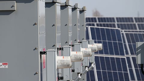 Row of industrial grey battery storage cabinets standing beside a large solar panel farm under clear sunlight.