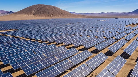 Aerial view of extensive rows of solar panel arrays spanning across a dry, desert landscape in Chile, with arid hills and mountains in the background under a clear blue sky.