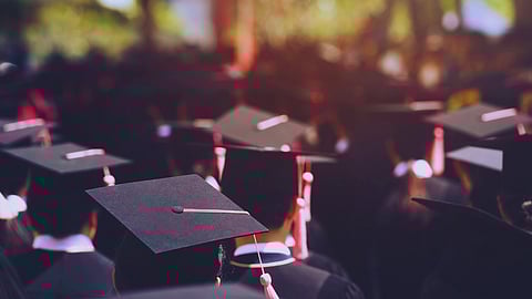 A crowd of graduates wearing black caps and gowns, adorned with pink tassels, is gathered outdoors. The mood is celebratory, with sunlight filtering through trees.