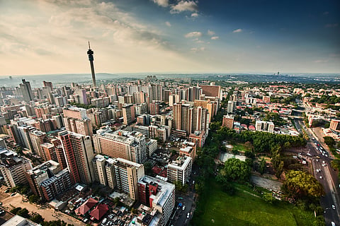 Aerial view of Johannesburg skyline with dense buildings, a tall communications tower, and green parks. The scene is under a blue sky with scattered clouds.