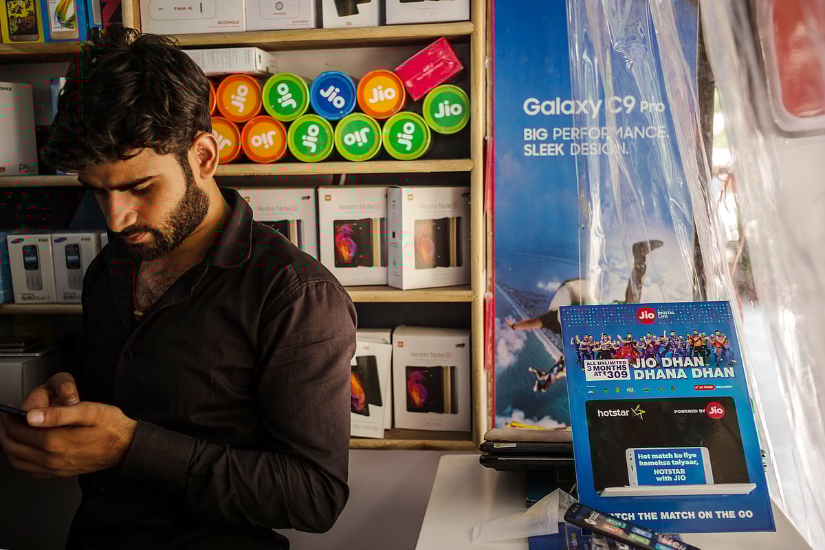 A vendor uses a smartphone while standing next to advertising for Reliance Jio Infocomm Ltd. at the Nehru Place IT Market in New Delhi. (Photographer: Sanjit Das/Bloomberg)