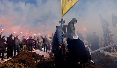 The image depicts a solemn military funeral ceremony taking place during winter in Ukraine.