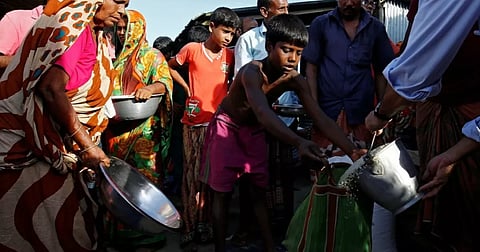 The image shows flood-affected villagers, including women and children, holding metal bowls and containers to collect relief food supplies distributed in Gaibandha, Bangladesh.