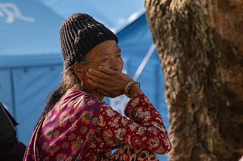 An elderly woman sits outside a temporary shelter, her hand resting against her face, as displaced families confront the uncertainty of life after the floods