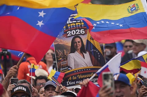 The image shows Venezuelan opposition leader and Nobel Peace Prize laureate María Corina Machado depicted on a campaign poster held up by demonstrators waving Venezuelan flags.