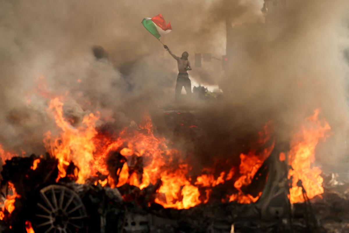 A man waves a Mexican flag as smoke and flames rise from a burning vehicle during a protest against federal immigration sweeps, near Los Angeles City Hall, in downtown Los Angeles, California, U.S. June 8, 2025.
