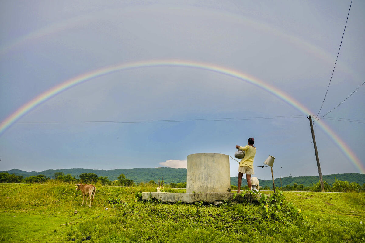 A woman fetches water from a well as a double rainbow appears in the sky after rainfall, in Kamrup