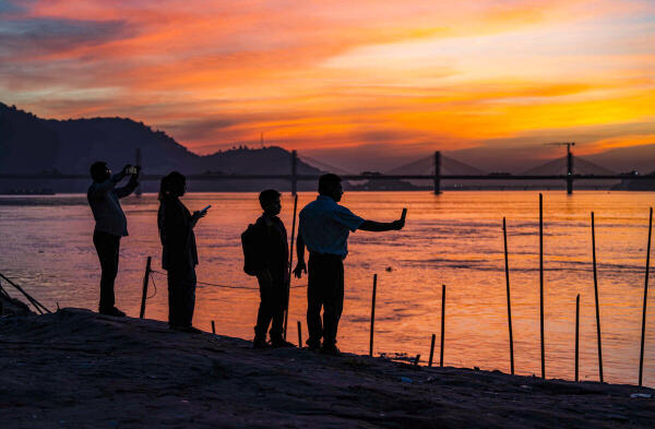 People during the sunset by the Brahmaputra riverside, capturing photos and moments against the scenic backdrop of a cable-stayed bridge, in Guwahati, Assam.