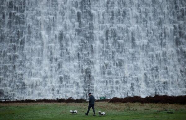 A man walks with his dogs in front of the wall of the Derwent Dam as water spills over the top following a prolonged period of rain, near Edale.