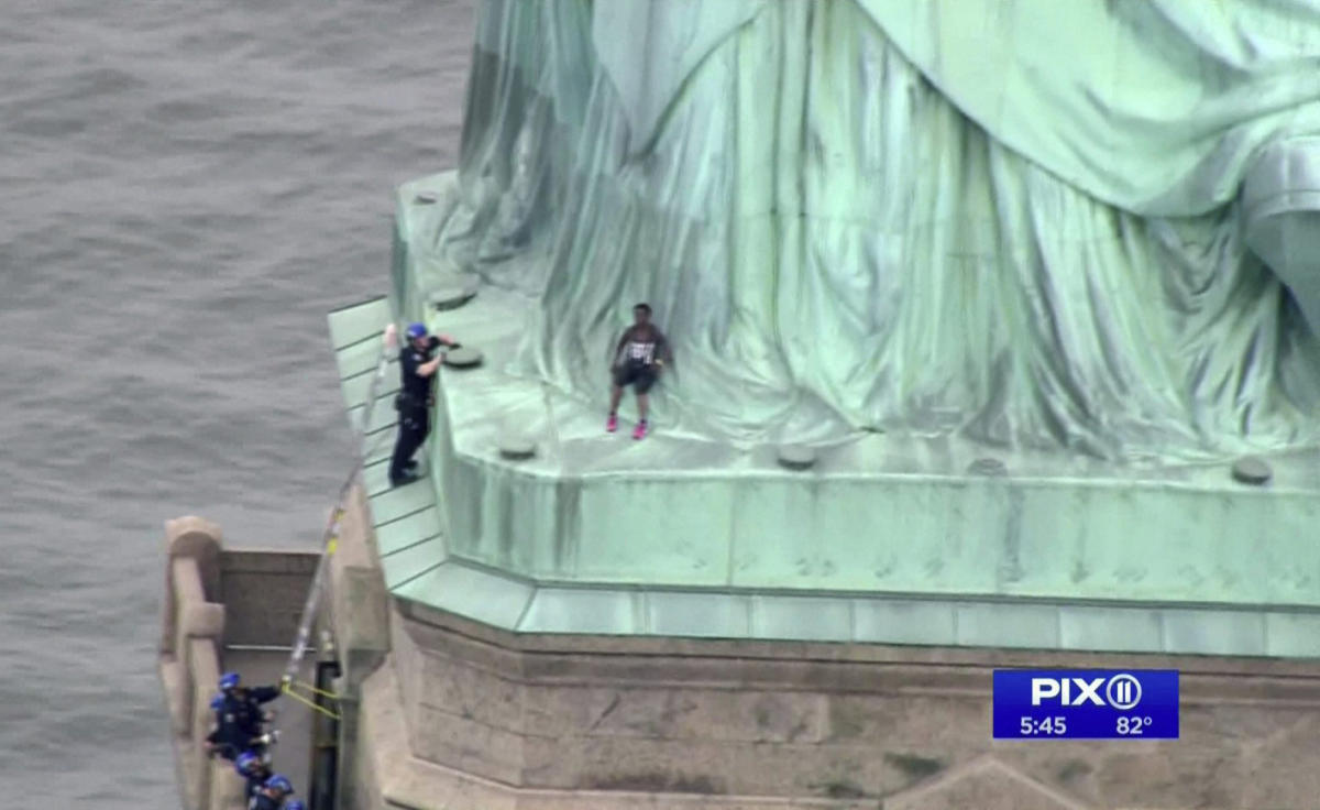 In this image made from video by PIX11, a person, center, leans against the robes of the Statue of Liberty on Liberty Island, as one of the police officers climbed up on a ladder to stand on a ledge nearby talking the climber into descending in New York, Wednesday, July 4, 2018. AP/PTI