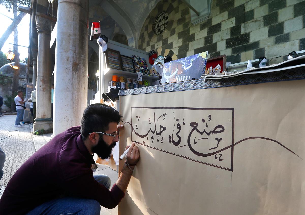 A merchant writes 'Made in Aleppo' on a stand in a trade fair dedicated to war-hit businesses from Aleppo looking to make a revival, in the Syrian capital Damascus' Tekkiyeh Sulimaniyeh complex. Ghazal is among 137 merchants from the northern province of Aleppo taking part in the state-sponsored week-long fair for small-scale businesses reeling from a nine-year conflict. Credit: AFP Photo