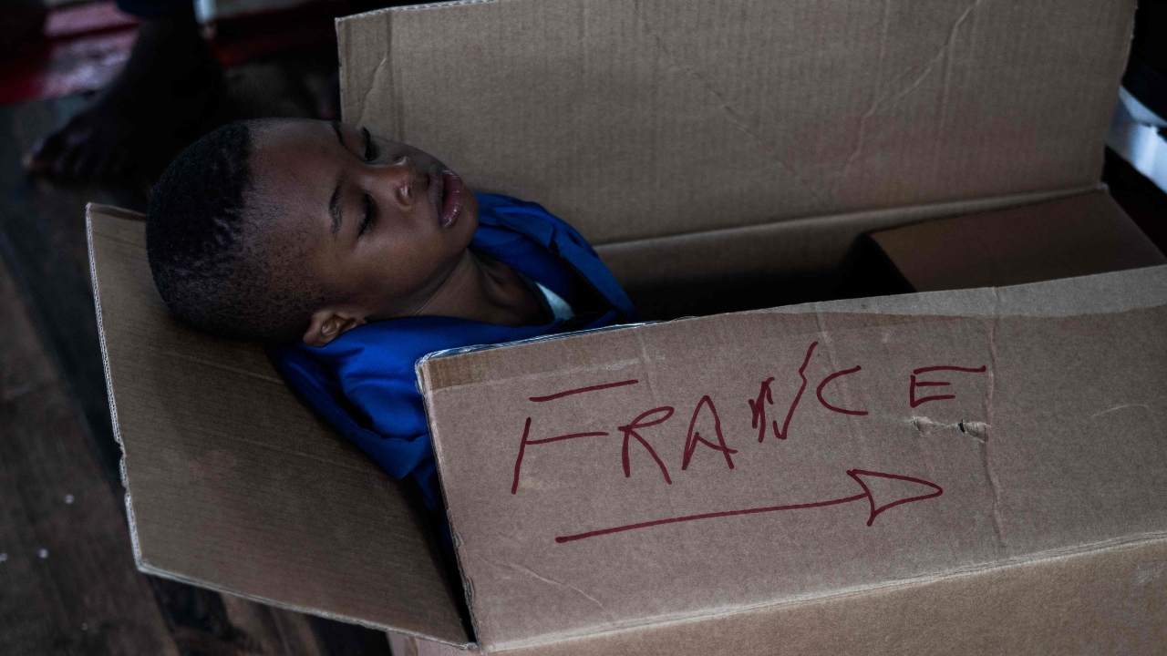 A migrant child sits in a cardboard box with the inscription