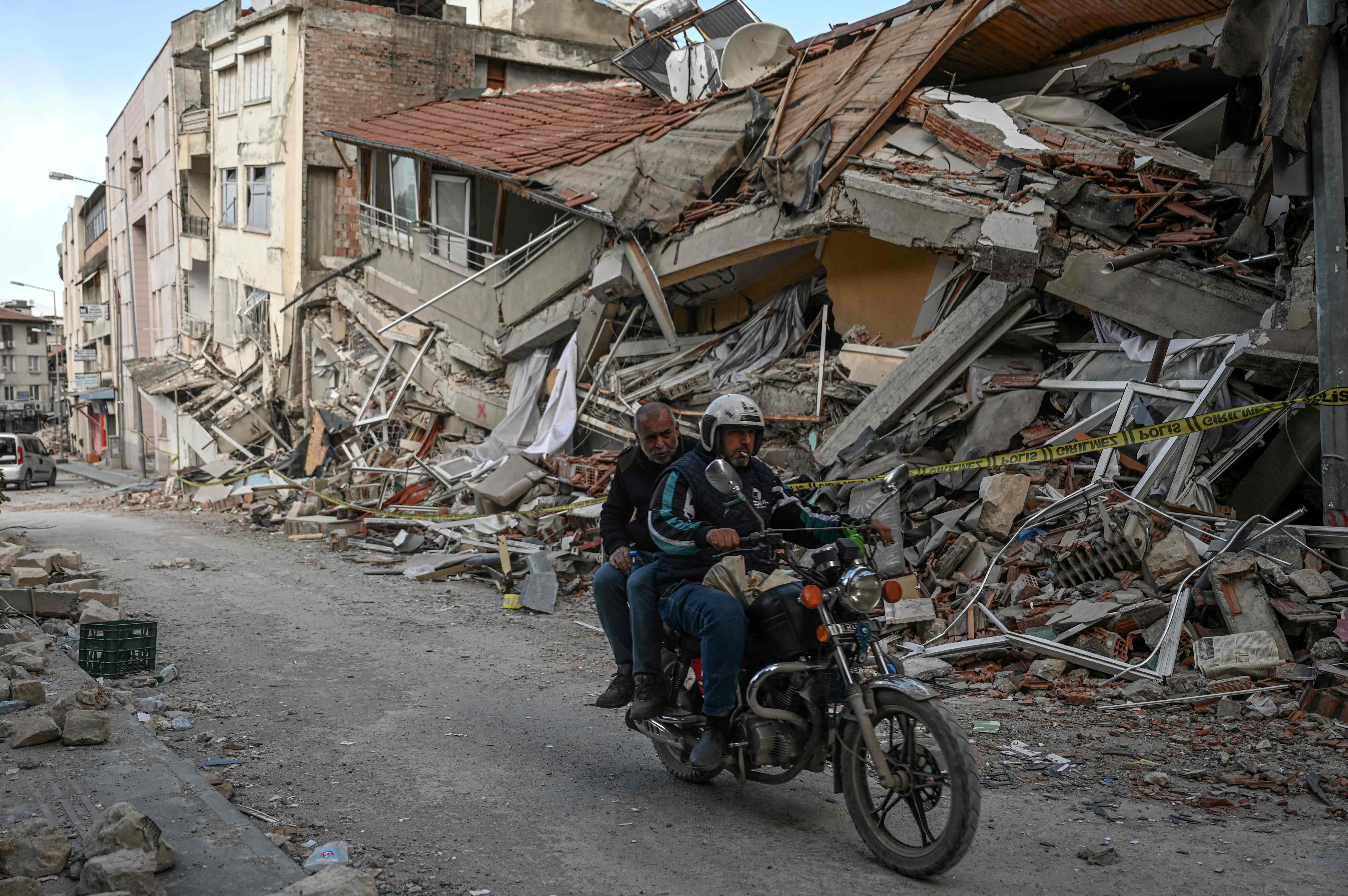 People ride a motorcycle in a street next to the rubble of collapsed buildings in Hatay. Credit: AFP Photo