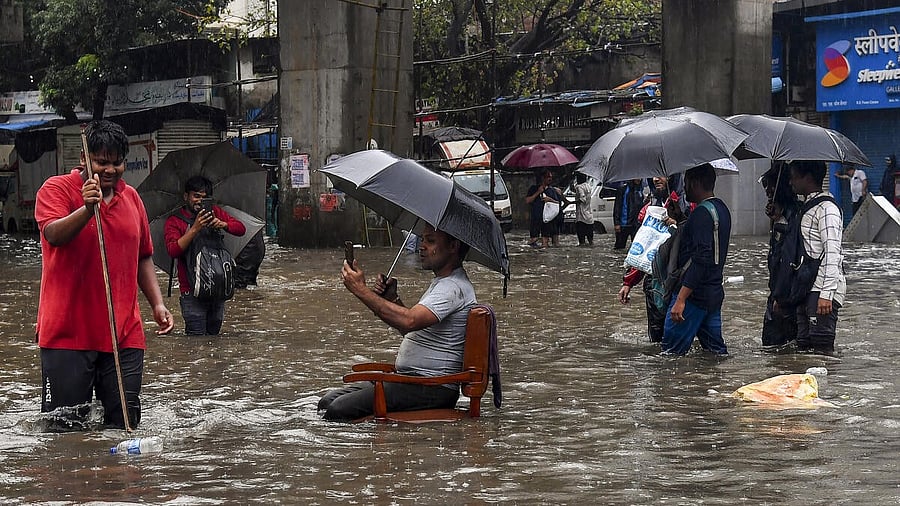 <div class="paragraphs"><p>A man sits on a chair as people wade through a waterlogged road amid rainfall, in Mumbai.</p></div>
