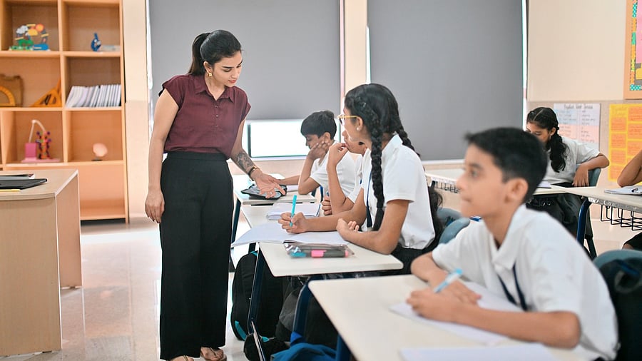 <div class="paragraphs"><p>Female school teacher bends over a student’s desk to give individual guidance while children in uniforms work on their notebooks, showing a supportive learning environment in a bright classroom. </p></div>