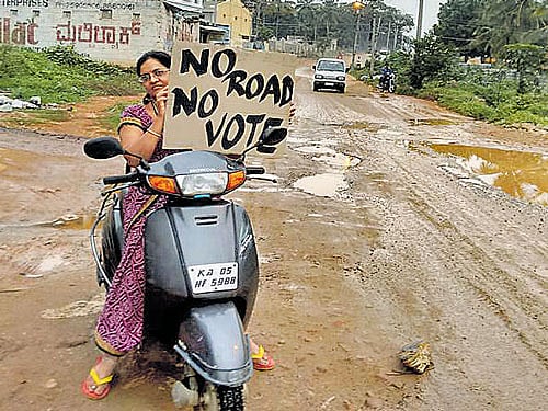 A resident holds a 'No Road No Vote' placard on a road in a bad condition at Rajarajeshwari Nagar. dh photo