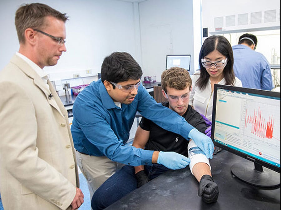 Graduate students Mengya Li and Nitin Muralidharan adjust the energy harvesting device on the arm of undergraduate Thomas Metke while Professor Cary Pint looks on. (John Russell / Vanderbilt)