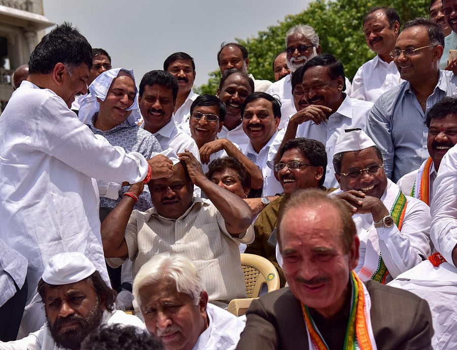 Congress leader D K Shivakumar puts a cap on the head of JD(S) state president H D Kumaraswamy as other Congress leaders look on during a joint protest at the Vidhana Soudha in Bengaluru on Thursday. dh photo