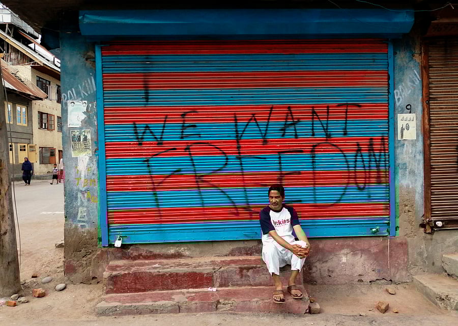  a man sits next to graffiti that reads "We Want Freedom" on a shuttered store in the Soura locality in Srinagar, during the lockdown. Photo by Jalees ANDRABI / AFP