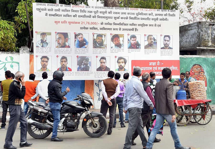 People walk past a poster displaying photographs of those who have been identified to pay the compensation for vandalizing public properties during protests against CAA. (PTI Photo)