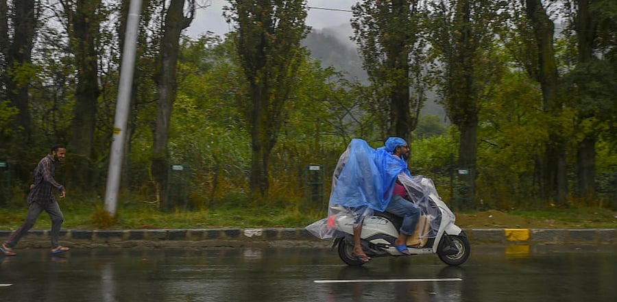 Men riding on a scooter cover themselves with a polythene sheet during heavy rain, in Srinagar. Credit: PTI