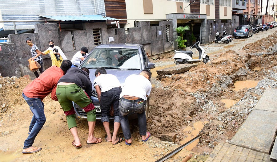 Residents try to push a car stuck on a muddy road at Nehru Nagar, Gandhinagar, following rainfall on Wednesday. DH PHOTO/B H SHIVAKUMAR