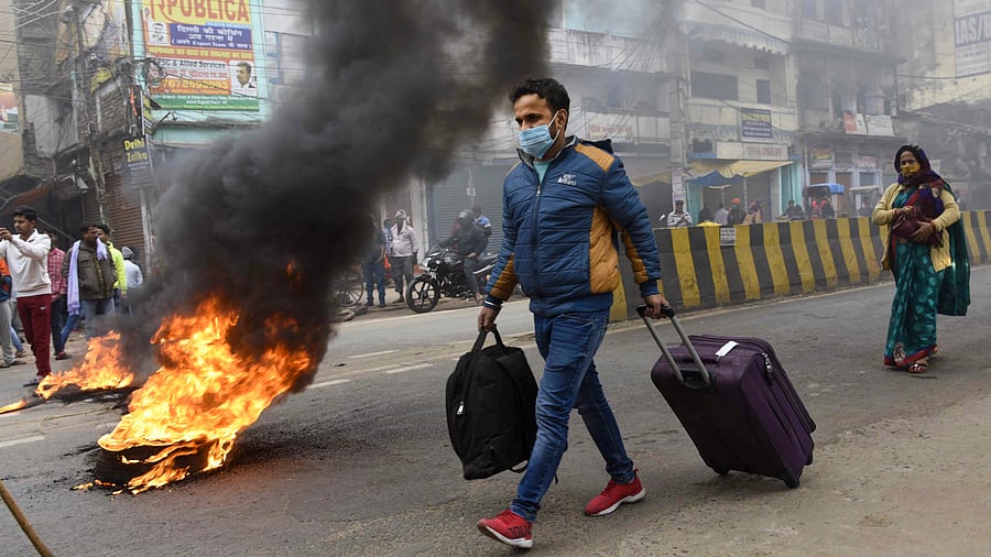 People with their luggage walk past a burning tyre, set ablaze by demonstrators in support of the nationwide strike called by agitating farmers against Centre's farm reform laws, in Patna. Credit: PTI Photo