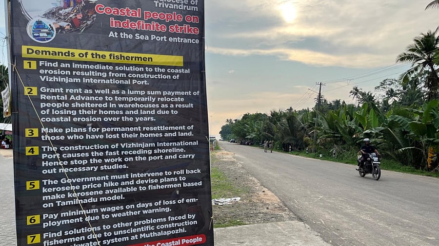 A motorcyclist moves past a placard featuring the demands of protesting fishermen near the entrance of the proposed Vizhinjam Port in Kerala. Credit: Reuters photo