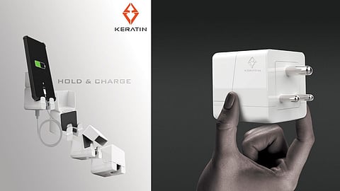 A sleek white mobile charger with dual functions is shown in this product image for the brand "KERATIN". The left side of the image demonstrates the charger acting as both a power adapter and a phone holder, with a phone charging while standing upright in the holder. The phrase "HOLD & CHARGE" is displayed near the top. The right side shows a hand holding the compact charger, highlighting its minimalist cube-like design and glossy finish. The KERATIN logo is visible on both the charger and the upper part of the image.
