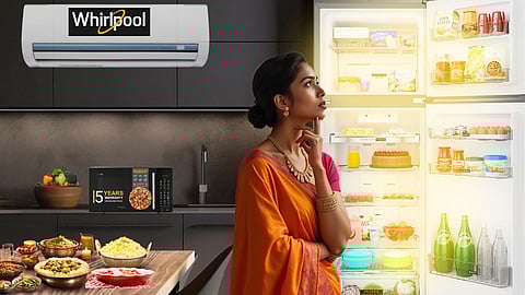 An Indian woman in an orange saree gazes thoughtfully at an open, brightly lit fridge in a modern kitchen with Whirlpool appliances.