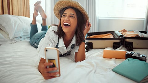 Smiling young woman in casual clothes and straw hat lying on a bed with smartphone, open suitcase, and travel accessories in a cosy homestay room