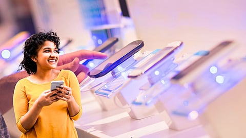 Smiling woman holding a smartphone in front of a display of new mobile phones at a brightly lit electronics store
