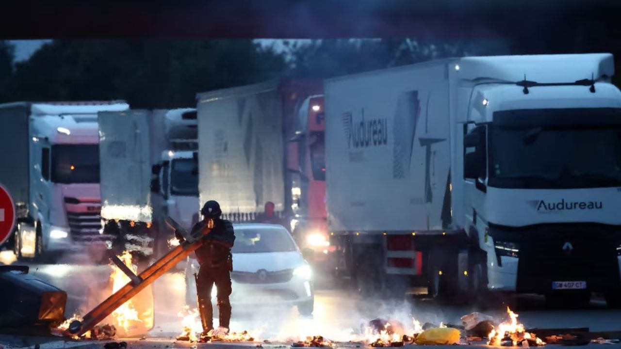 Police officer standing in front of burning barricades on a road while trucks and cars wait behind during a protest or blockade in France.
