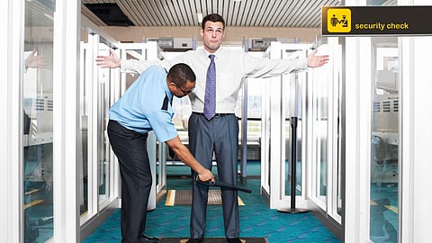 Security officer conducting a body scan with a handheld detector on a man in formal attire during an airport security check, with a sign reading ‘security check’ visible in the background