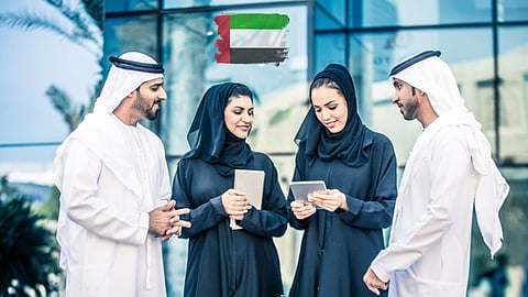 Group of Emirati professionals in traditional attire discussing work outdoors, with UAE flag displayed above