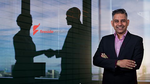 A professional man in a suit smiling confidently in front of a glass wall with the Fireside Ventures logo, while two businesspeople are seen shaking hands in reflection, symbolising investment and partnerships