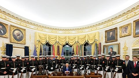 Donald Trump seated at the Resolute Desk in the Oval Office, surrounded by a large group of uniformed US Marines, with portraits and gold curtains in the background