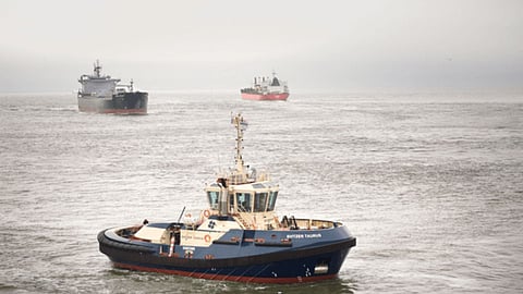 A small blue-and-white tugboat named Svitzer Taurus sails in the foreground on a grey, misty sea, with two larger cargo ships visible in the distance. The overcast sky and muted tones create a calm maritime atmosphere