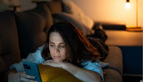 Young serious sad woman lying on sofa and looking at smartphone screen at night time at home