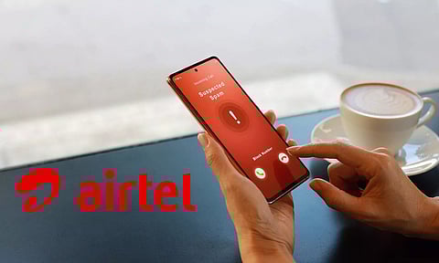 Person holding a smartphone showing an incoming call screen marked ‘Suspected Spam’ with options to accept or reject the call. Airtel logo is displayed prominently on the left. A cup of coffee is placed on the table in the background
