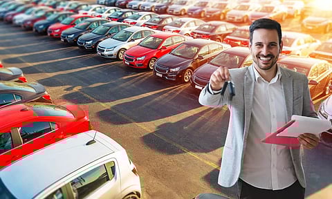 Smiling car salesman holding keys and documents in front of a large row of parked cars at a dealership lot during sunset