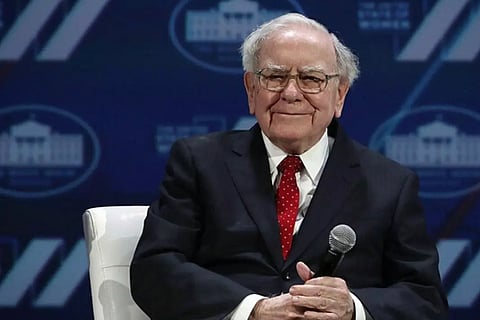 An elderly man in a dark suit, white shirt, and red polka dot tie sitting on stage with a microphone in hand, smiling at the audience. A blue backdrop with white logos is visible behind him