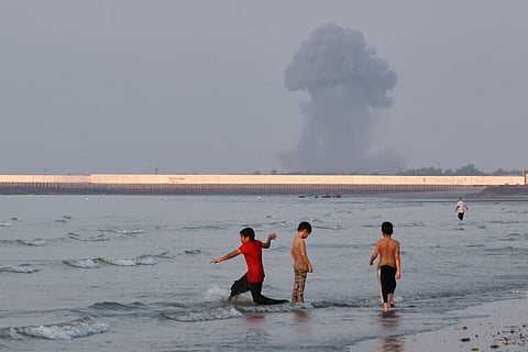 Three boys play in the shallow waters of the Strait of Hormuz, as a plume of smoke rises from an explosion in the background, off Bandar Abbas, Iran, Monday, July 13, 2026.