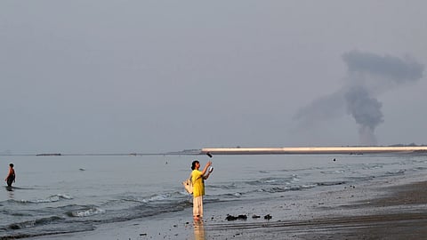 A woman stands at the water's edge along the Strait of Hormuz as a plume of smoke rises in the background following an explosion, off Bandar Abbas, Iran