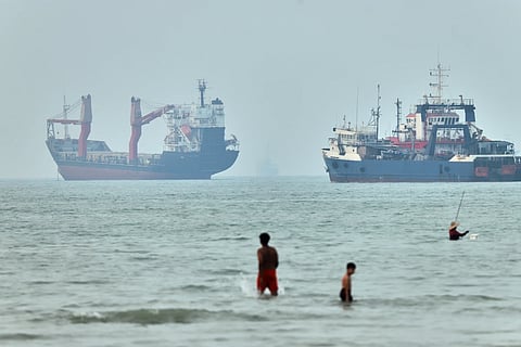 Children wade in the water with cargo ships at anchor in the background and a fisherman nearby, in the Strait of Hormuz off Bandar Abbas, Iran,