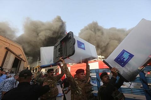 Security forces carry ballot boxes as smoke rises from a storage site in Baghdad