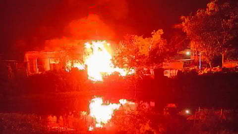 A massive fire engulfs a building at night, casting a bright orange glow that reflects off the surface of a nearby body of water